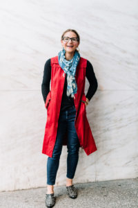 woman smiling wearing a red vest open with denim jeans that are slim fitting and a blue denim scarf against a white marble wall
